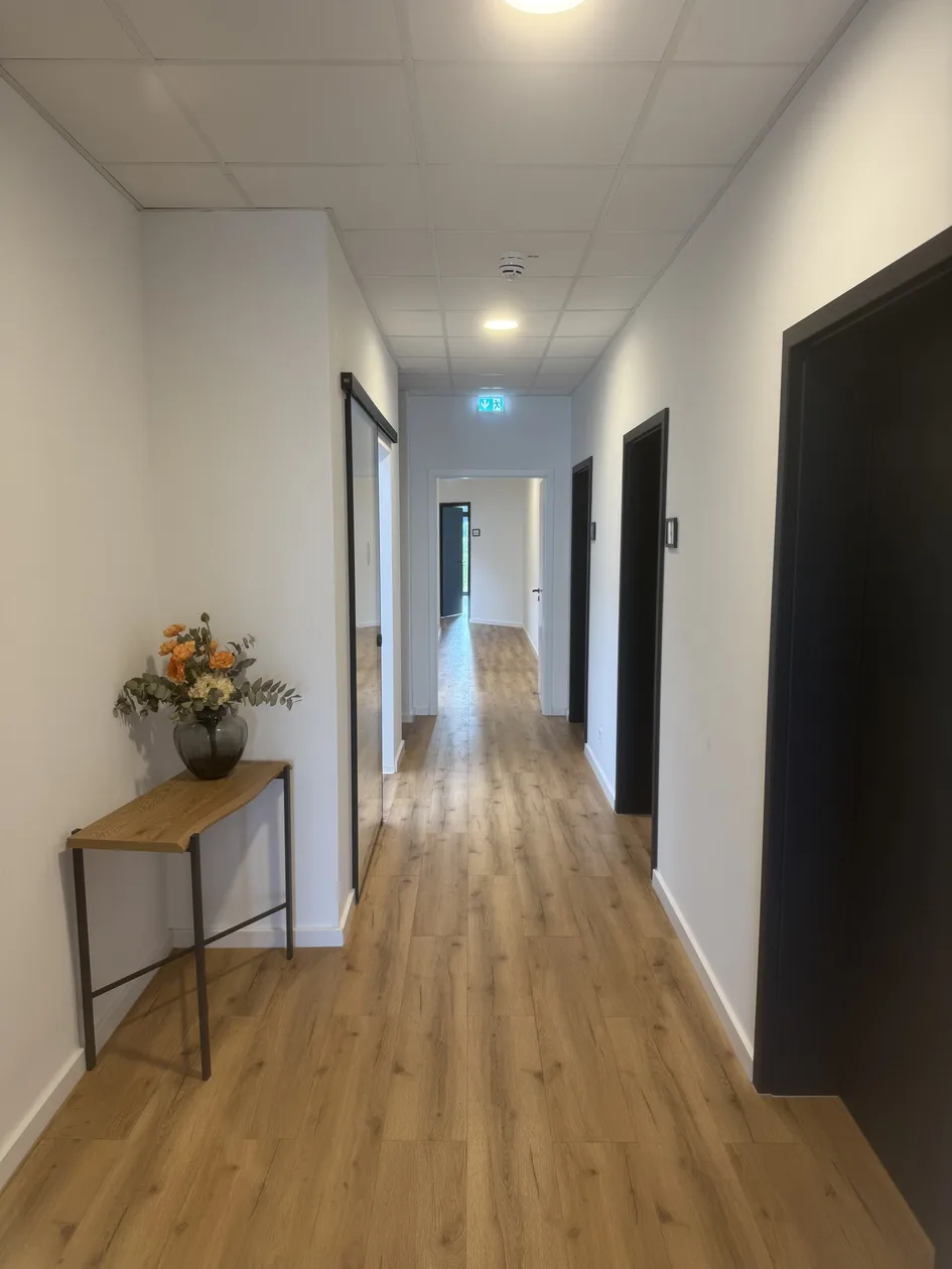 Heller Flur mit Holzboden, Behandlungsraumtüren und Beistelltisch mit Blumen